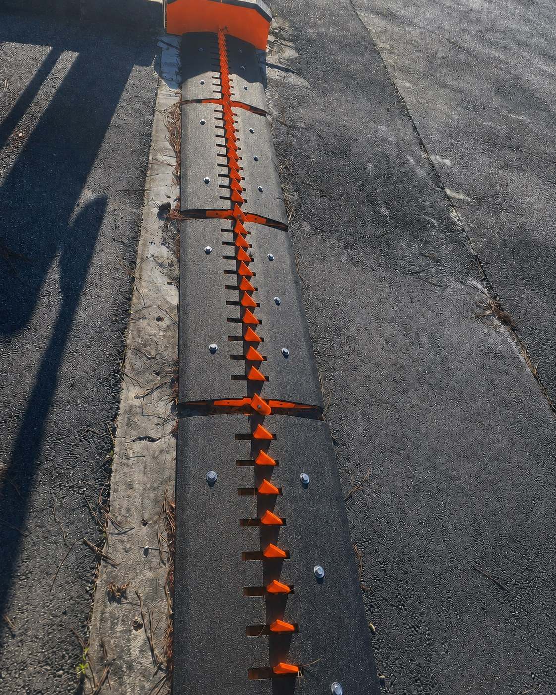 Overhead daylight view of a surface-mounted traffic spike run with orange blades on a black median housing, harsh sun and long shadows
